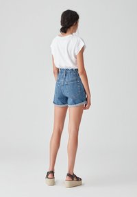 Mujer de pie de espaldas, vistiendo una camiseta blanca de manga corta, pantalones cortos vaqueros de tiro alto y sandalias de cuña negras sobre un fondo liso.