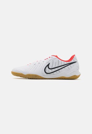 Biały sportowy but na halę z czarnym logo Nike, czerwoną wyściółką i gumową podeszwą, pokazany z boku na białym tle.