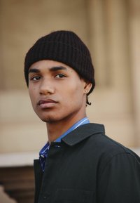 Sort ribbet strikket beanie, blåstribet skjorte under en sort jakke. Nærbillede af en ung mand med kort hår og en stud-ørering.