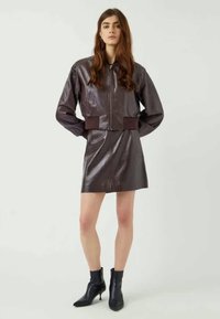 Bruine cropped jas van imitatie leer met ritssluiting, met brede mouwen en een bijpassende bruine mini-rok, gecombineerd met zwarte enkellaarsjes.