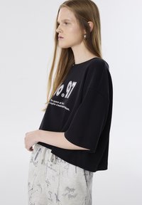 Zwart cropped T-shirt met korte mouwen, voorzien van een witte grafische print. Gecombineerd met lichtgrijze joggingbroek met een textuurpatroon.