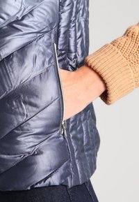 Puffjacka med en glänsande marinblå yta, som har chevron-sömmar och en ficka med dragkedja. Bruna stickade manschetter förhöjer designen.