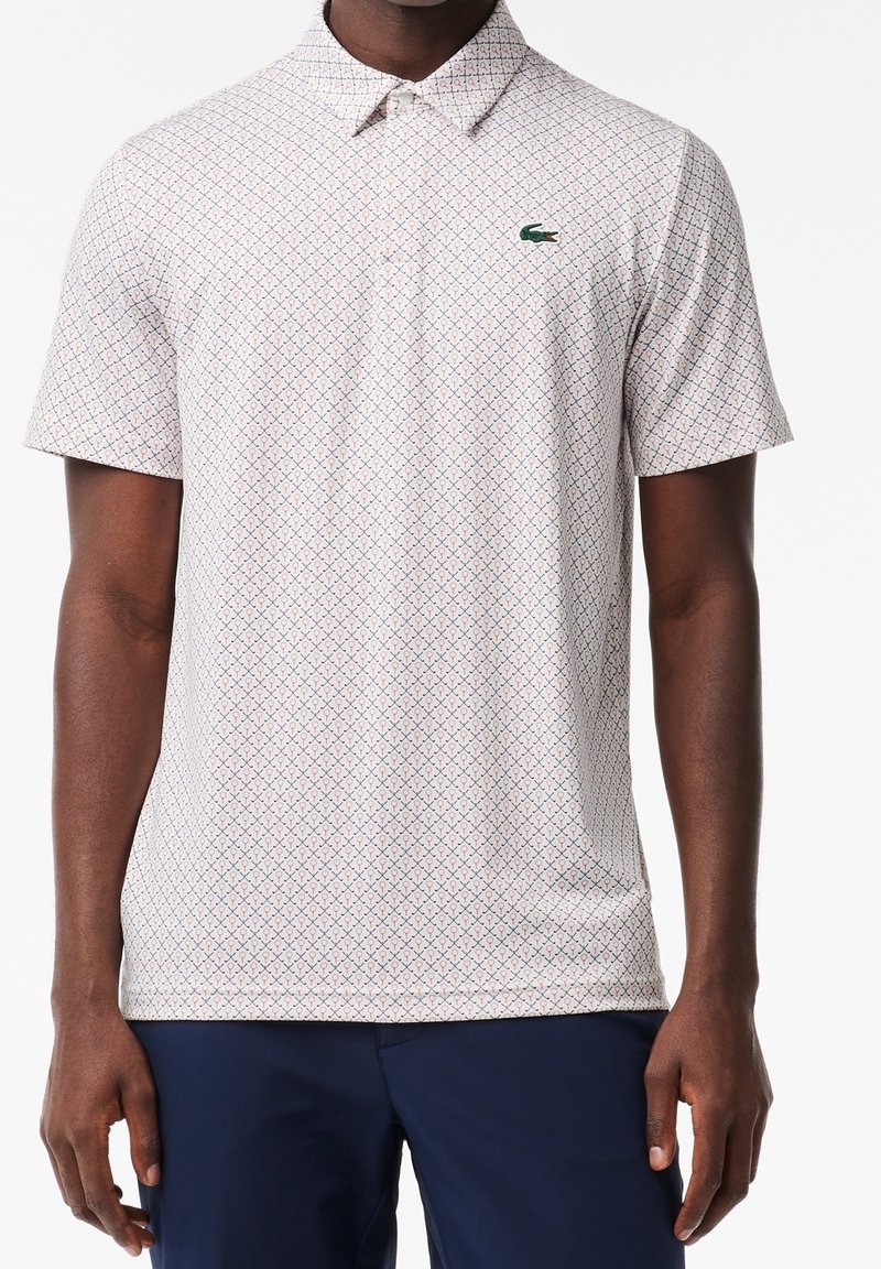Homme portant un polo à manches courtes de couleur claire avec un petit motif géométrique et un logo de crocodile vert, associé à un pantalon bleu marine foncé.