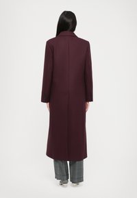 Langer, tief burgunderfarbener Mantel mit Reverskragen, Rückenschlitz und Knopfakzenten an den Ärmeln, kombiniert mit karierter Hose und Schuhen.