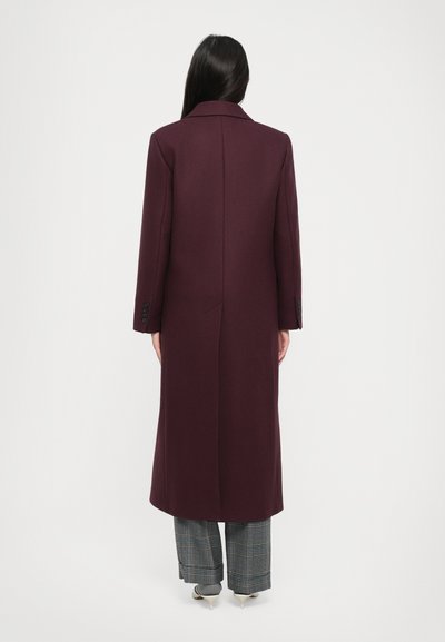 Pitkä, syvä viininpunainen takki, jossa on viistetyt kaulukset, selkäventtiili ja napit hihassa, yhdistettynä ruudullisiin housuihin ja kenkiin.