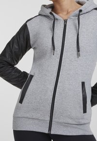 Persona vistiendo una sudadera con cremallera gris con mangas de cuero sintético acolchado negro y ribetes de bolsillo negros, con las manos en las caderas.