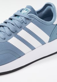 Ljusblå sneakers med tre vita ränder, texturerad syntetöverdel, rund tå, snörningsdesign och svart gummisula.