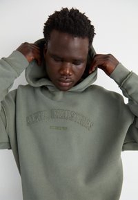 Hoodie vert olive avec une poche kangourou et le logo "ALPHA INDUSTRIES" brodé, en tissu épais et avec une capuche à cordon ajustable.