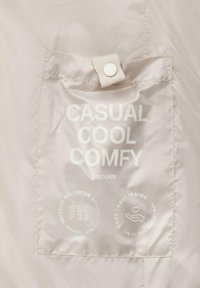 Etykieta z recyklingowego poliestru w jasnym beżu z białym napisem "CASUAL COOL COMFY" oraz symbolami pielęgnacyjnymi. Widoczne detale guzika i szwów.