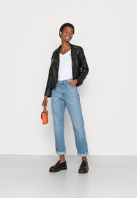 Chaqueta de cuero negra, camiseta blanca de cuello en V, jeans claros arremangados, zapatos negros, sosteniendo una bolsa naranja. Diseño simple y ajuste casual.