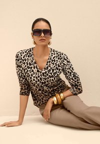 Frau mit übergroßen dunklen Sonnenbrillen, Leopardenmuster-Oberteil, beigen Hosen, großen goldenen Ohrringen und klobigen goldenen Armbändern, sitzt vor einem beige Hintergrund.