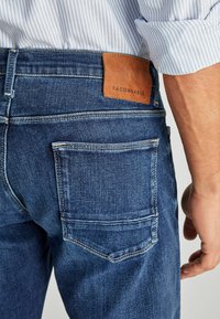 Jeans de mezclilla azul oscuro con un parche de cuero etiquetado "FACONNABLE" en la cintura, con costuras en contraste y un diseño de bolsillo trasero.
