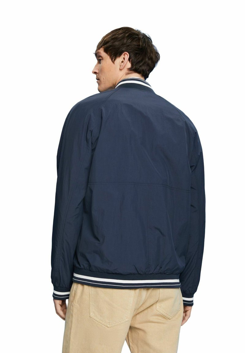 Blouson de pilote bleu marine avec un col et des poignets côtelés, présentant une texture lisse et un ourlet élastique. Porté avec un pantalon de couleur claire.