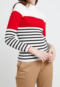 Top rouge et blanc côtelé avec des rayures horizontales noires, col montant, et des accents rouges festonnés sur les manches, associé à un pantalon beige.