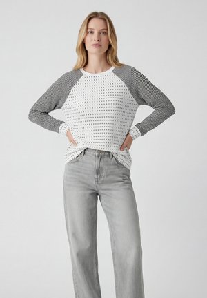 Blondynka stoi, ubrana w biały sweter w czarne kropki i paski na rękawach oraz jasnoszare dżinsy z rękami w kieszeniach.