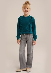 Zelena rebrasta majica s dolgimi rokavi in naborki, v kombinaciji z zbledelimi sivo širokimi hlačami in svetlimi čevlji. Preprost, sproščen outfit.