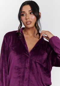Plush sammet violett skjorta med krage, knäppning fram och bröstficka. Slät struktur, avslappnad passform, synliga sömnadsdetaljer.