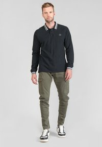 Zwarte lange mouwen polo met grijs gestreepte kraag, gecombineerd met olijfgroene cargobroek en zwarte sneakers met witte zolen en accenten.