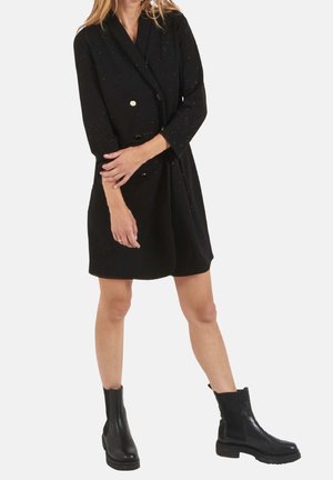 Zwarte dubbelgeknoopte jurk met gouden knopen, driekwartmouwen en een licht losse pasvorm, gedragen met zwarte enkellaarzen. Subtiele spikkels zichtbaar.