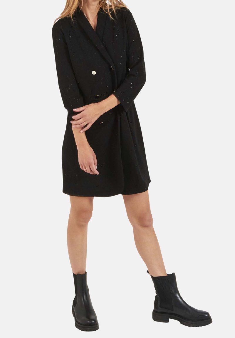 Robe noire à double boutonnage avec des boutons dorés, manches trois-quarts et coupe légèrement ample, portée avec des bottines noires. Légères taches discrètes visibles.