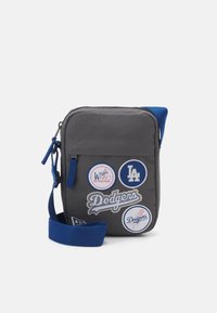 Borsa a tracolla grigia in tessuto con dettagli blu, decorata con vari patch a tema Dodgers. Include una chiusura con zip e una tracolla regolabile.