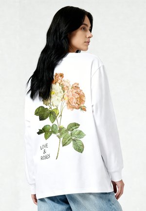 Frau mit langen dunklen Haaren, die ein weißes Langarmshirt mit großem Blumenmuster und dem Schriftzug „LOVE & ROSES“ auf dem Rücken trägt.
