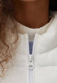 White quilted jacket with a high collar, featuring a front zipper and ribbed white fabric at the neckline and underlayer.