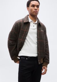 Καφέ καρό μπουφάν bomber με κουμπωτό μπροστινό μέρος και ριμπ μανσέτες, συνδυασμένο με λευκό πόλο πουκάμισο και μαύρο παντελόνι. Διακριτό λογότυπο γνωστής μάρκας στο μπουφάν.