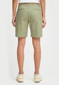 Person iført lysegrønne shorts til knærne, hvite sneakers og en hvit topp, som står med ryggen til kameraet mot en ensfarget bakgrunn.