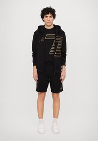 Black zip-up hoodie with gold graphic design, black t-shirt, black shorts, and white sneakers. Simple, casual athletic wear.