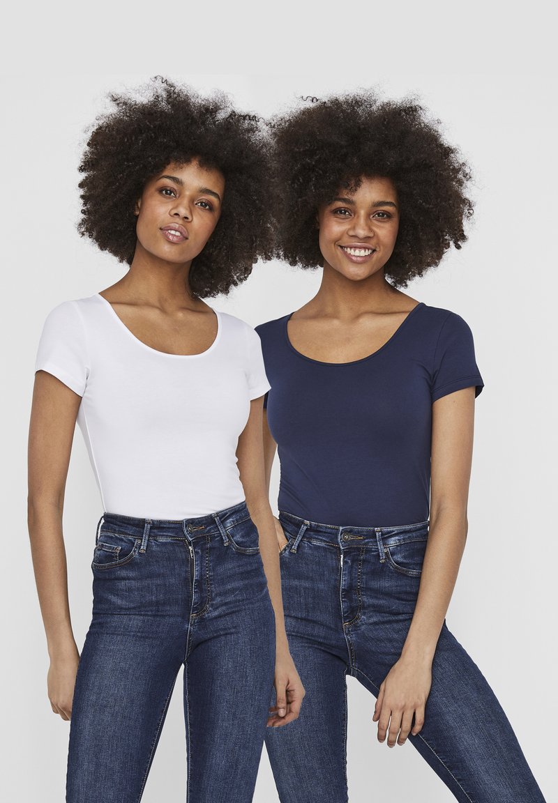 Deux femmes portant des hauts ajustés ; l'une est blanche, l'autre est bleu marine. Toutes deux ont des manches courtes et sont associées à des jeans en denim bleu.