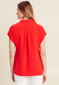Polo shirt rosso a manica corta con colletto, tessuto strutturato, dettaglio di piega posteriore e orlo curvo, abbinato a pantaloni bianchi aderenti.