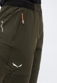 Κοντινή λήψη των αθλητικών παντελονιών σε ελαιοπράσινο χρώμα με τσέπη στο μηρό που κλείνει με φερμουάρ, πορτοκαλί τράβηγμα φερμουάρ και λευκό λογότυπο κοντά στην τσέπη στο αριστερό πόδι.