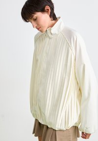 Giacca plissettata color crema chiaro con colletto alto, chiusura con zip e orlo elasticizzato; presenta un tessuto liscio e dettagli ai lati.