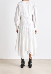Abito bianco con maniche lunghe, dettaglio ad occhio di pernice e orlo asimmetrico. Indossato con stivaletti neri alla caviglia, che mostrano un tessuto texturizzato.