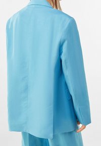 Osoba v světle modrém saku a odpovídajících kalhotách, zobrazená zezadu, s jednou rukou viditelnou u jejich strany.