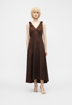 Kvinne som står iført en brun ermeløs midi-kjole med V-hals, rysjer på stropper og hvite åpne hæler på en ensfarget bakgrunn.