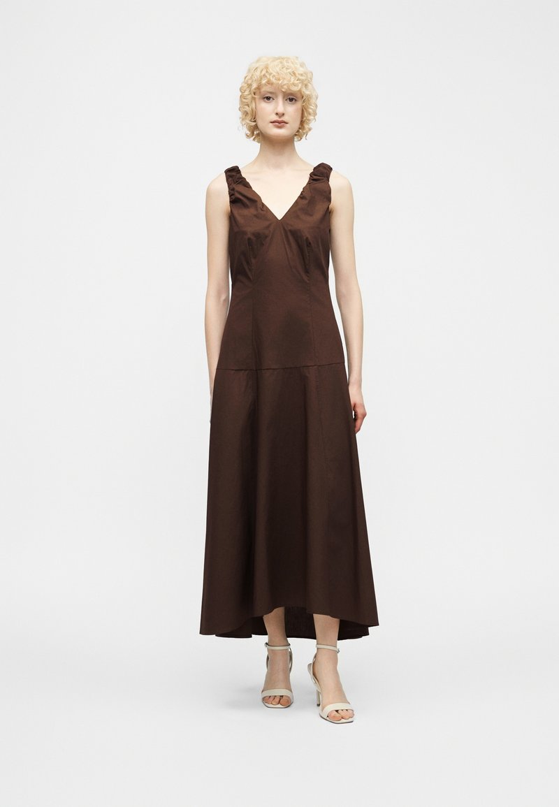 Γυναίκα που στέκεται φορώντας ένα καφέ μίντι φόρεμα χωρίς μανίκια με V λαιμόκοψη, με ραμμένες βολάν στους ιμάντες και λευκά πέδιλα ανοιχτής μύτης, σε απλό φόντο.