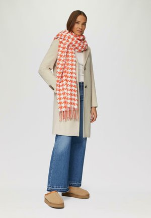 Écharpe à motif pied-de-poule en orange et blanc, en tissu doux, avec des bords frangés. Portée avec un léger manteau et un jean à jambes larges.