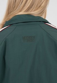 Personne blonde vue de dos, portant une veste vert foncé avec des rayures blanches et rouges sur les épaules et "MODERN YUPPIES" brodé au dos.