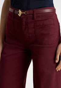 Tumši sarkanas bikses ar gludu tekstūru, kurās ir priekšējās kabatas un kontrastējošs brūns josta ar zelta sprādzes detaļu.
