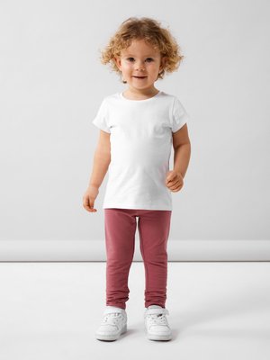 Dijete nosi bijelu majicu s kratkim rukavima i ružičaste tajice, u kombinaciji s bijelim tenisicama. Ima kovrčavu kosu i neutralnu pozadinu.