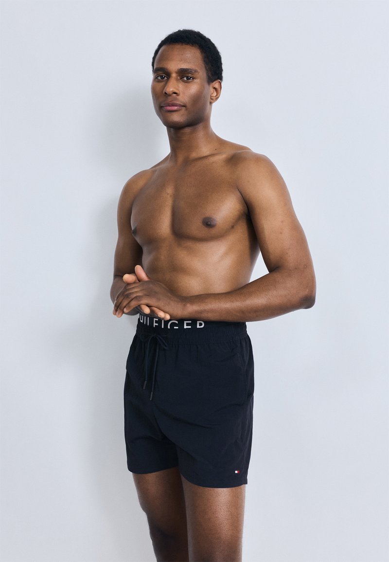 Μαύρο μαγιό σορτς με ελαστική μέση και ρυθμιζόμενο κορδόνι, με λογότυπο στη ζώνη και κόκκινες λεπτομέρειες. Ομαλή υφή.