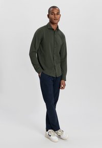 Cămașă button-up de un verde închis, cu mâneci lungi, pantaloni bleumarin și adidași albi. Cămașa are un guler standard și un închizător frontal.