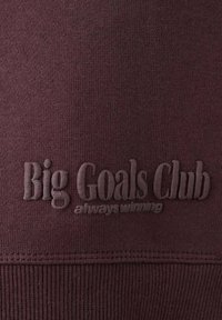 Tmavě červená látka mikiny s žebrovanou texturou. Obsahuje vyražený text "Big Goals Club" a "always winning" ve stejné barvě pro decentní značení.