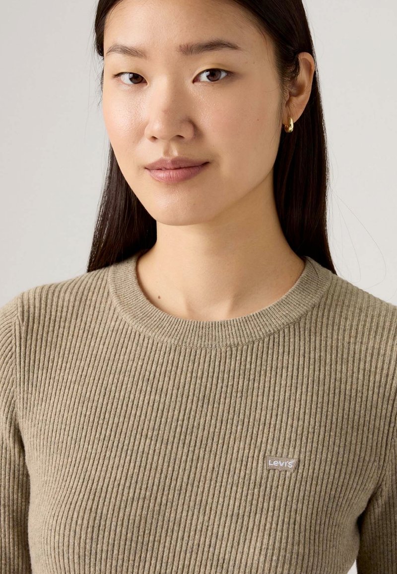 Giovane donna con capelli scuri e lisci indossa un maglione beige a coste con una piccola etichetta del marchio Levi's e un orecchino a cerchio dorato.