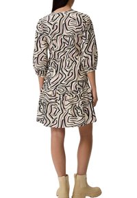 Musterkleid mit abstraktem Design in Schwarz, Weiß und Pink. Lange Ärmel, geraffte Taille, gestufter Rock mit knielangem Saum. Getragen mit beigen Knöchelstiefeln.