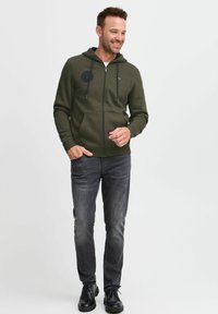 Sudadera con cremallera de color verde oscuro con detalles en negro y cordones, combinada con jeans grises y zapatos negros. La tela parece suave y texturizada.