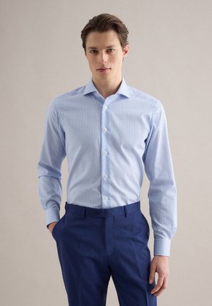 Junger Mann trägt ein hellblau-weiß gestreiftes Hemd, das in dunkelblaue Anzughose gesteckt ist, und steht vor einem schlichten Hintergrund.