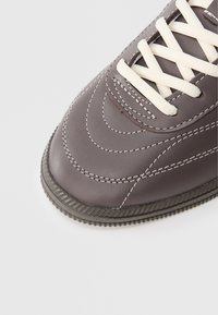 Καφέ μύτη αθλητικού παπουτσιού με λευκά κορδόνια, λεπτομερείς λευκές ραφές και υφή στο καουτσούκ της σόλας, πάνω σε απλό φωτεινό φόντο.